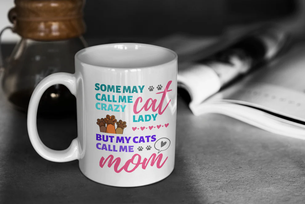 White mug with colorful text 'SOME MAY CALL ME CRAZY CAT LADY BUT MY CATS CALL ME MOM' on a dark surface.