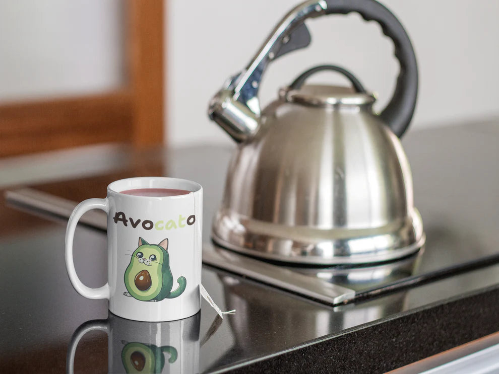 Mug with avocado design and cat illustration next to a silver kettle on a reflective surface.
