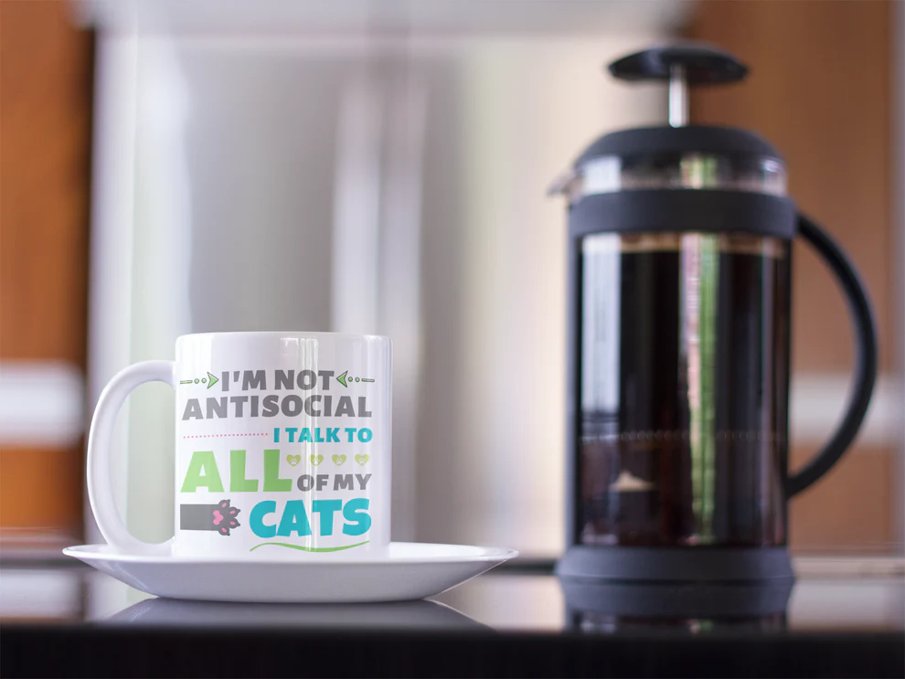 Mug with humorous text about cats on a kitchen counter with a French press in the background