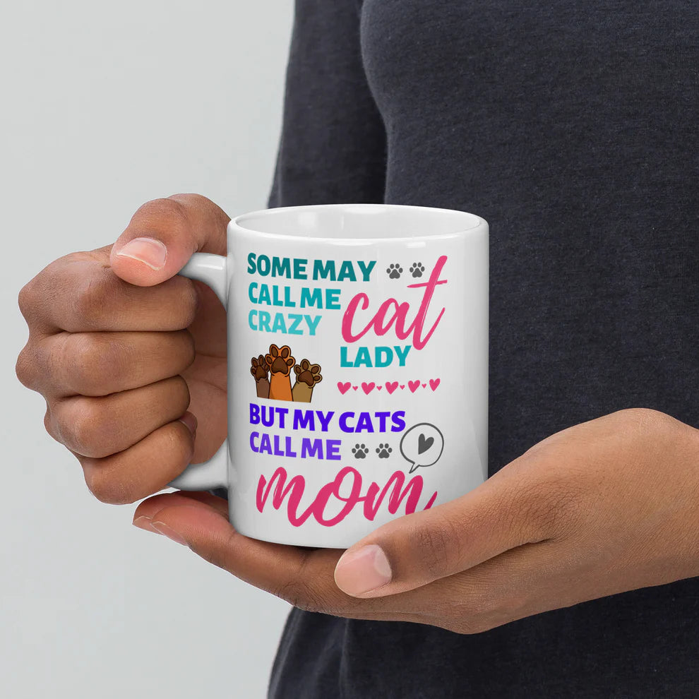Person holding a mug with text 'Some may call me crazy cat lady but my cats call me mom' on a plain background